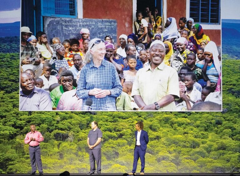 Three men presenting onstage with a photo of Jane Goodall in a crowd in the background Three men presenting onstage with a photo of Jane Goodall in a crowd in the background