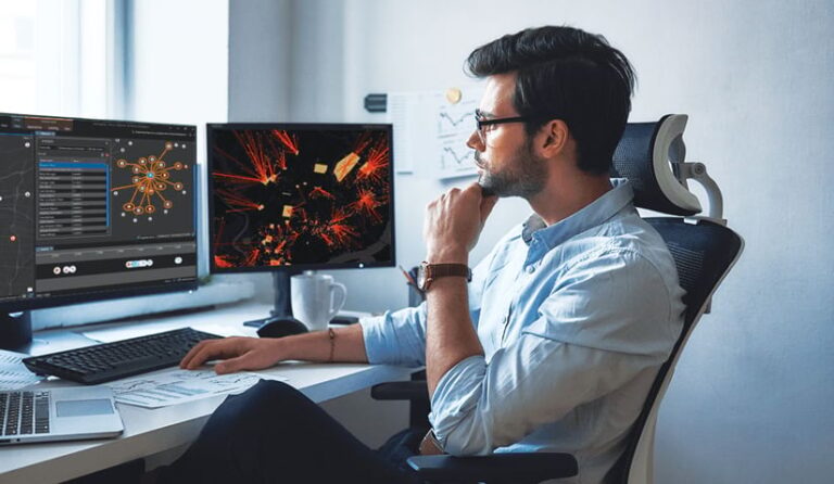 A man sitting at a desk looking at two monitors with maps and data on them A man sitting at a desk looking at two monitors with maps and data on them