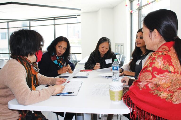 Five women sitting at a table, talking, and looking at papers