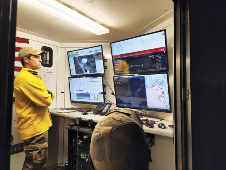 Two people in a portable building watching four monitors with maps, video feeds, and stats on them Two people in a portable building watching four monitors with maps, video feeds, and stats on them