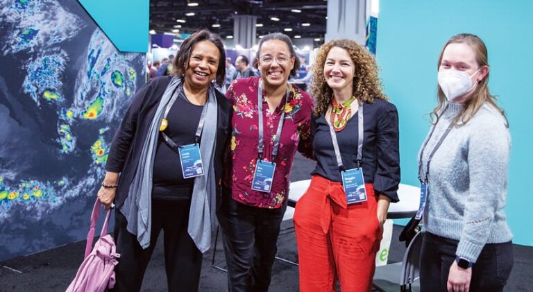 Four women posing for a photo in front of displays at a conference Four women posing for a photo in front of displays at a conference