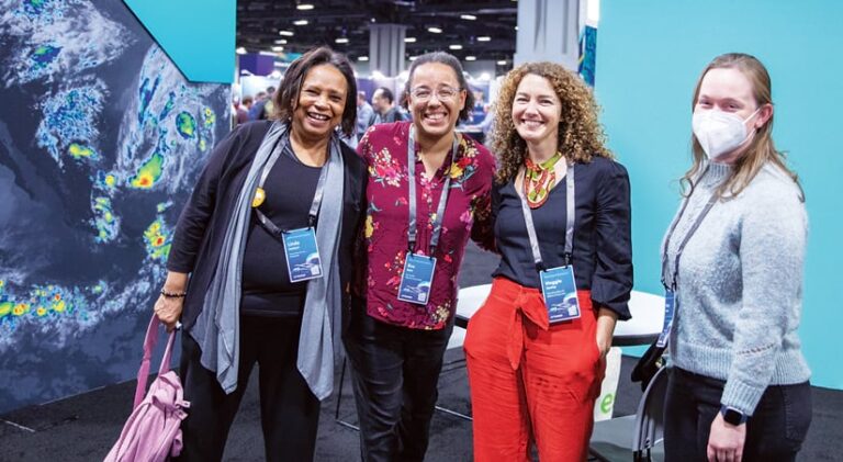 Four women posing for a photo in front of displays at a conference