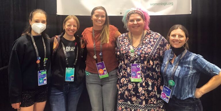 Five women posing for a photo in front of a Women in GIS sign Five women posing for a photo in front of a Women in GIS sign