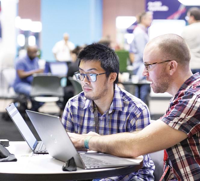 Two men sitting at a table, talking while looking and pointing at their laptops