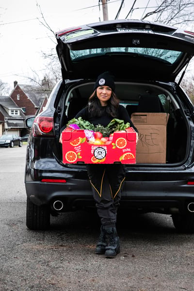 A woman standing at the back of her car holding a box full of fresh vegetables