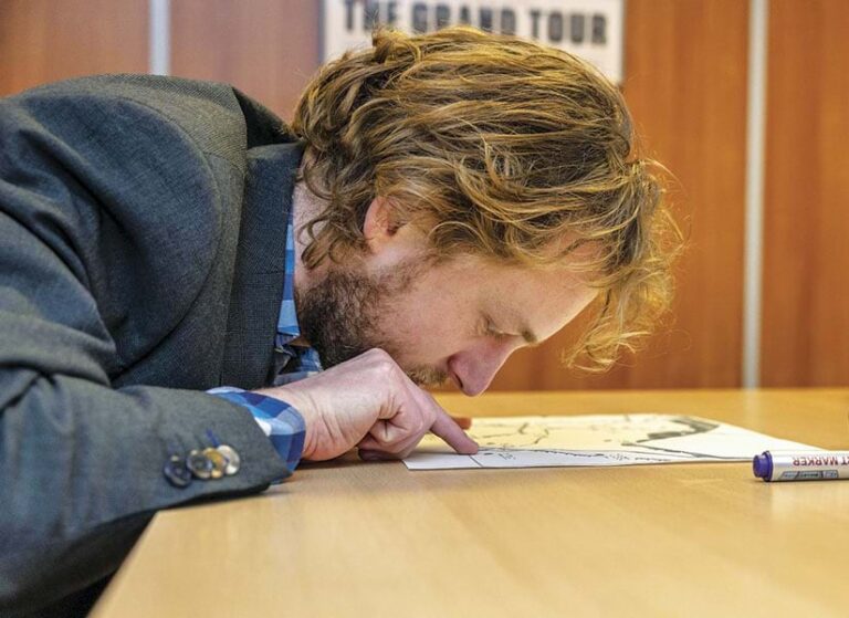 A man leaning over a table, touching a tactile map with his fingers and looking very closely at the map