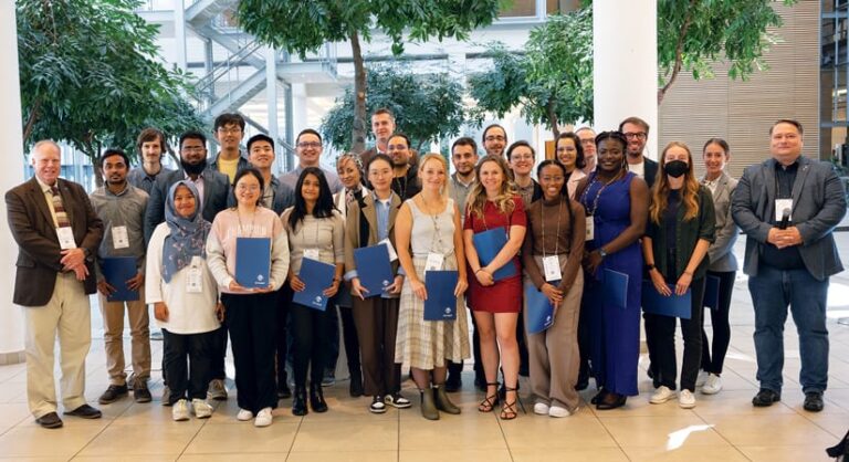 In the atrium area of a building, about 25 people, consisting mostly of young adults, pose for a group photo. Many have conference tags around their necks and hold blue folders.