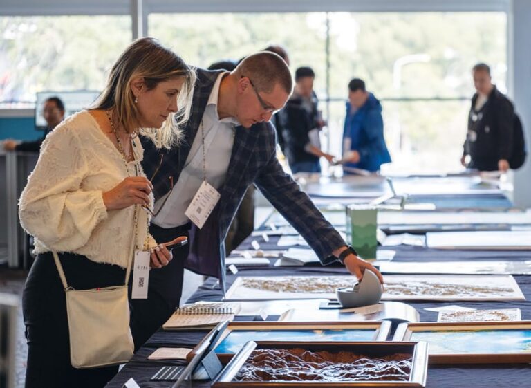 A few conference attendees peruse exhibits that lie on a table. A wooded area is visible through windows at the back of the room.