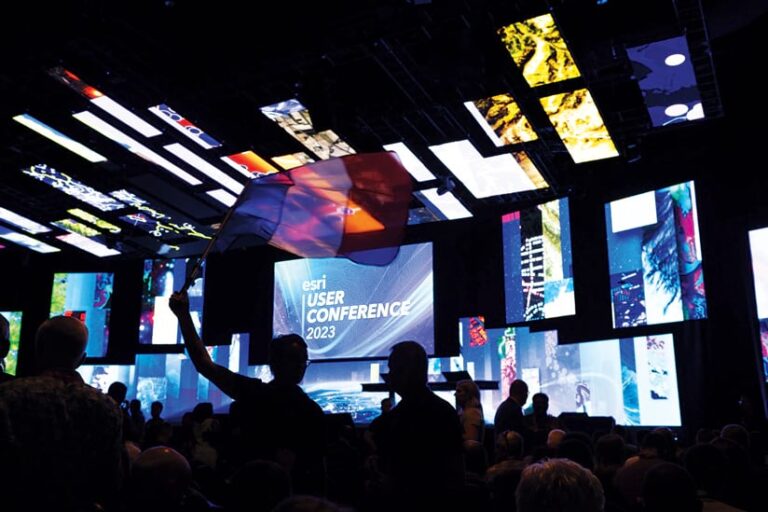 The Plenary stage in the background, showing colorful screens that display images and “Esri User Conference 2023,” and shadows of people in the foreground, with someone holding a French flag
