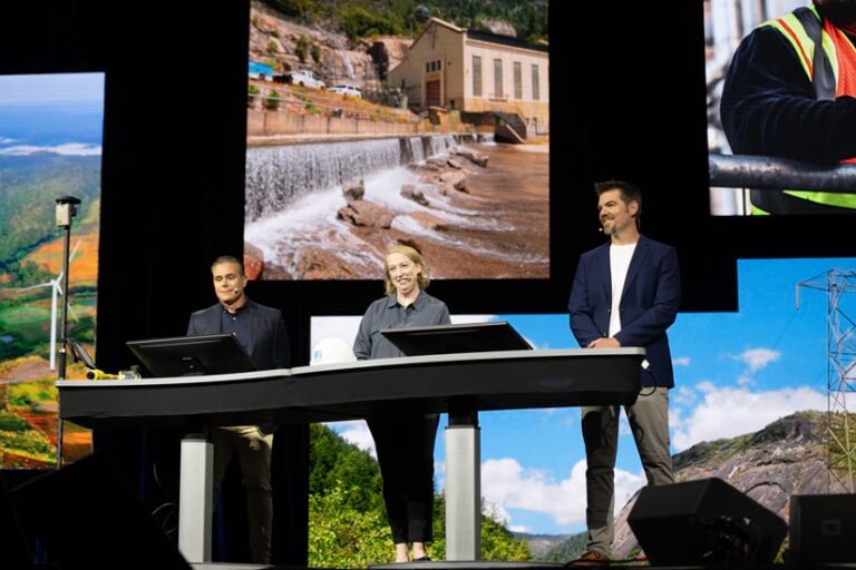 Three people standing on the Plenary stage behind a table with computer screens and in front of a big screen with images on it