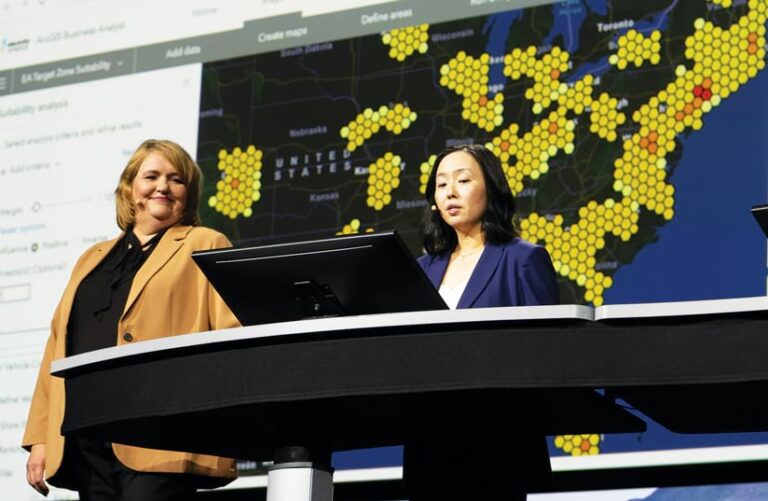 Two women standing on the Plenary stage in front of a big screen with a hexagonal map on it, with one woman standing behind a computer screen and the other one looking at her