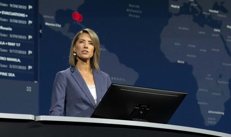 A woman speaking from behind a low computer screen while standing in front of a dark map of the world