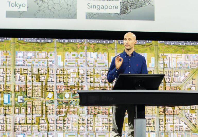 A man speaking on-stage, standing behind a table with a computer on it and in front of a giant screen with an aerial map on it