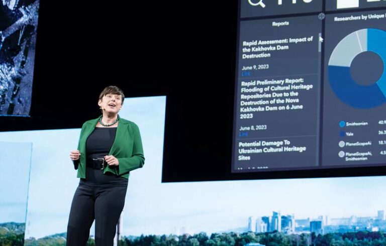 A woman in a green jacket speaking on-stage while standing in front of a big screen showing a dashboard