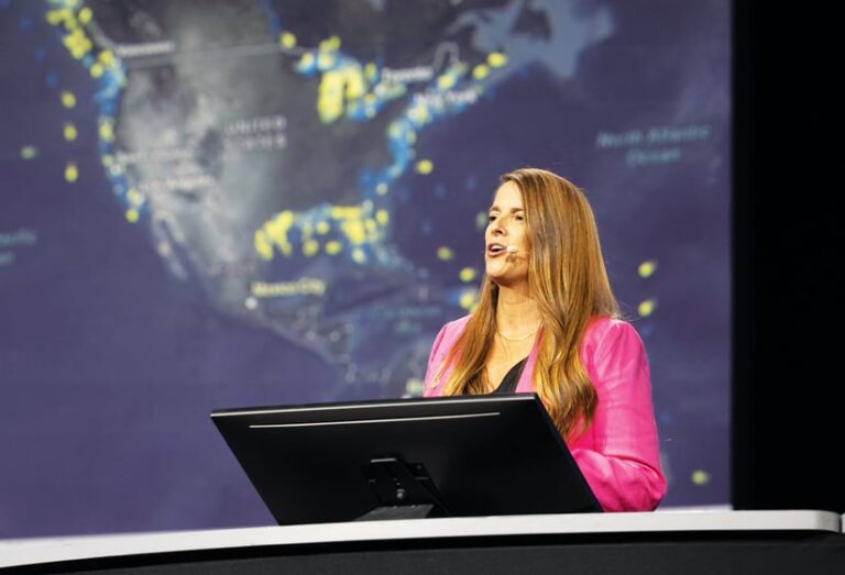A woman in a pink jacket speaking to the audience from behind a computer while standing in front of a map of North America