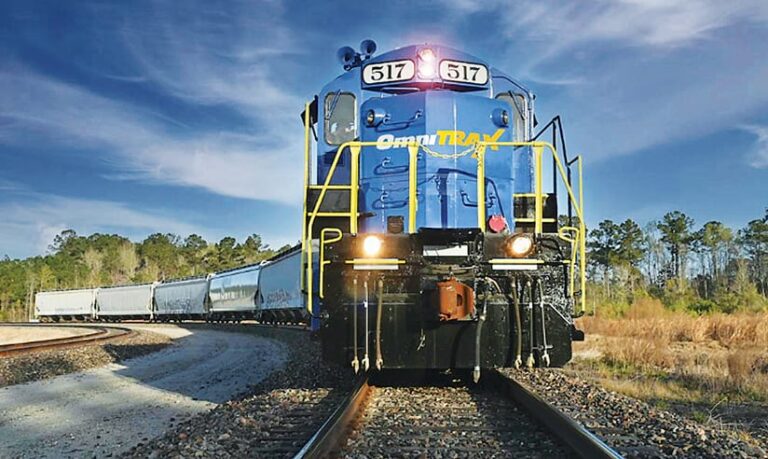 A train with at least six sections rounds a bend in a wooded and grassy area. The word OmniTRAX appears on the front of the train.