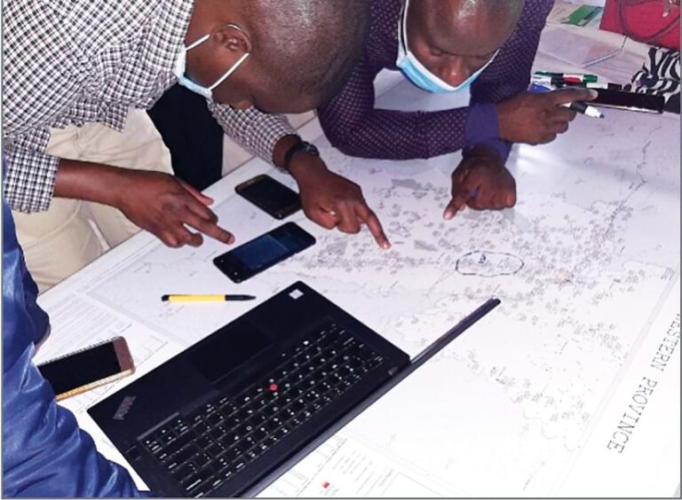 Two people in masks leaning over a laptop and a map, pointing at different places on the map