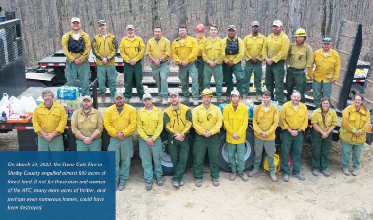 Twenty-three AFC workers pose on and next to a flatbed truck parked on the edge of a forested area.