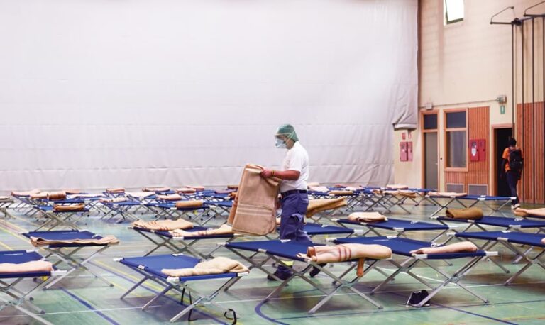 A health worker wearing a mask and hair net puts blankets on cots in an emergency shelter