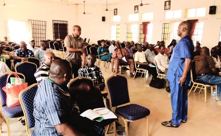 A person dressed in a blue shirt and pants addresses a room with mostly seated individuals.