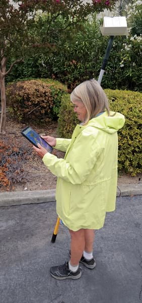 A person wearing a light yellow hooded raincoat carries a long tool and views a computer tablet.