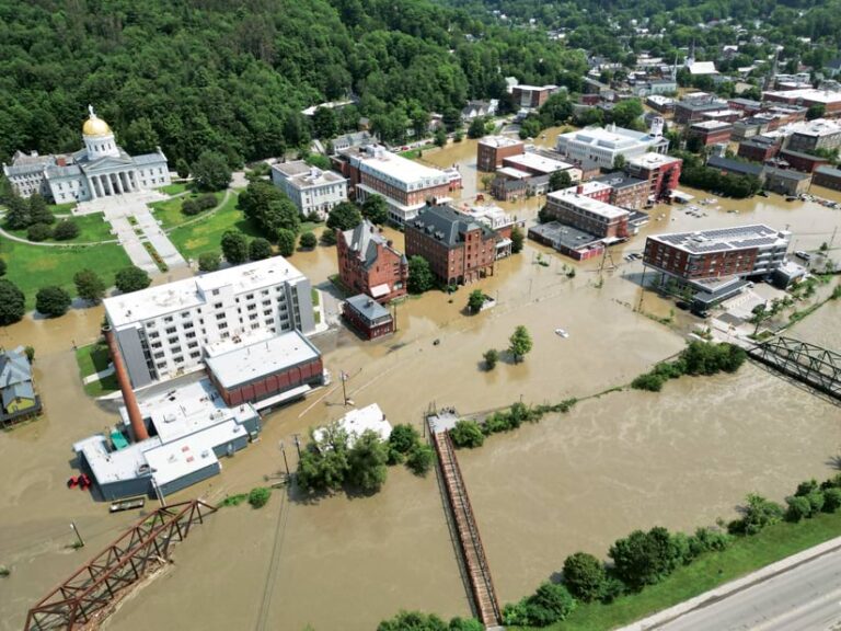 Floodwaters surround buildings in an urban area with a forested area in the background.