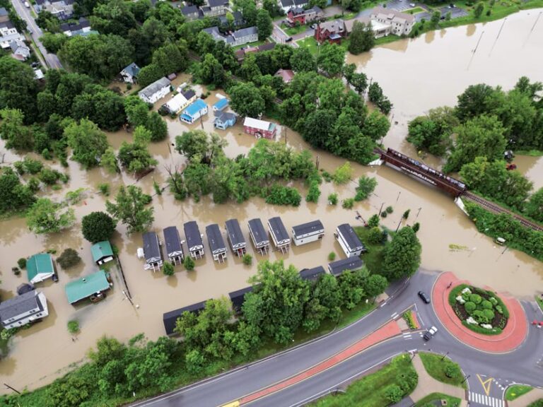 Floodwaters surround buildings and trees and cover part of a roadway.