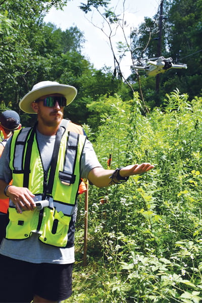 An individual wearing a wide-brimmed hat, sunglasses and a yellow vest holds an outstretched hand under a hovering drone.