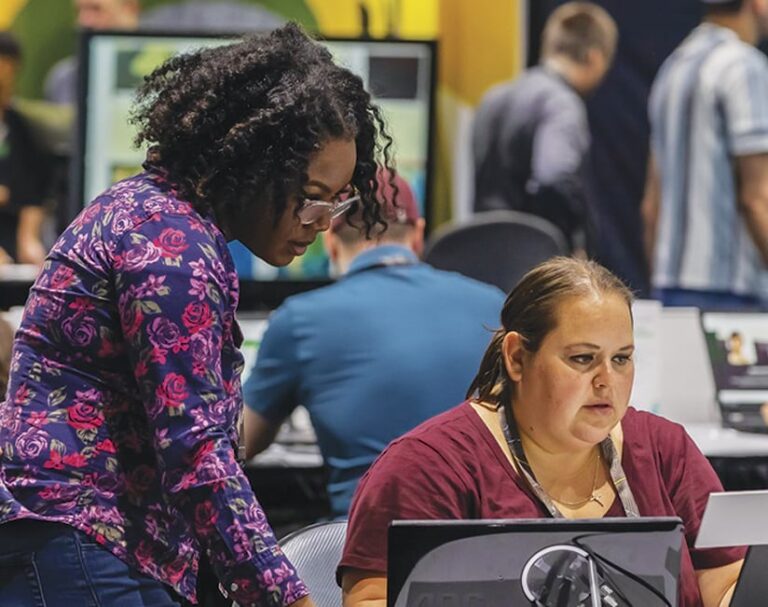 In a room with many computers and people, two individuals look at a computer.