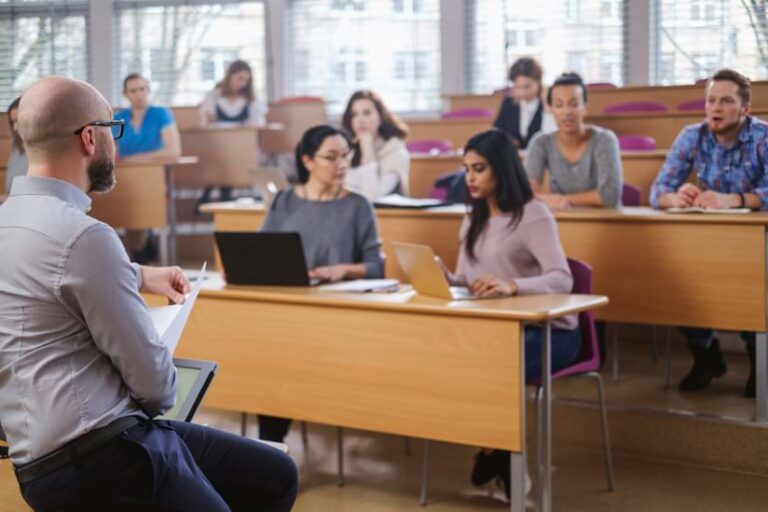 A professor stands in a tiered lecture hall addressing students working at individual desks. The students, who represent diverse ethnicities, are focused on laptops and notebooks. A professor stands in a tiered lecture hall addressing students working at individual desks. The students, who represent diverse ethnicities, are focused on laptops and notebooks.