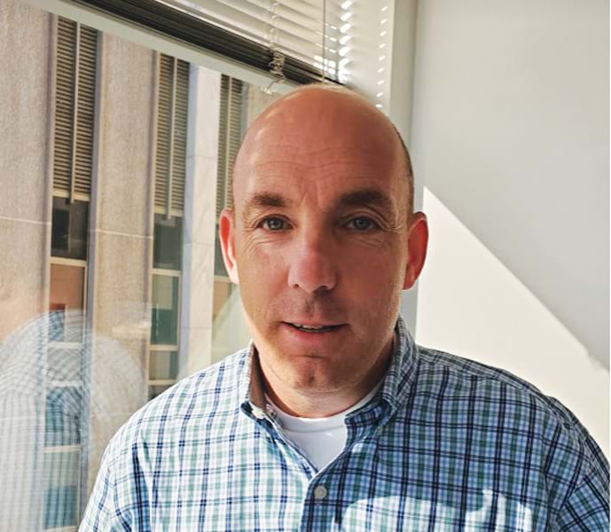 Headshot of a man with short hair and a blue and green plaid shirt, smiling in front of a window with blinds. Headshot of a man with short hair and a blue and green plaid shirt, smiling in front of a window with blinds.