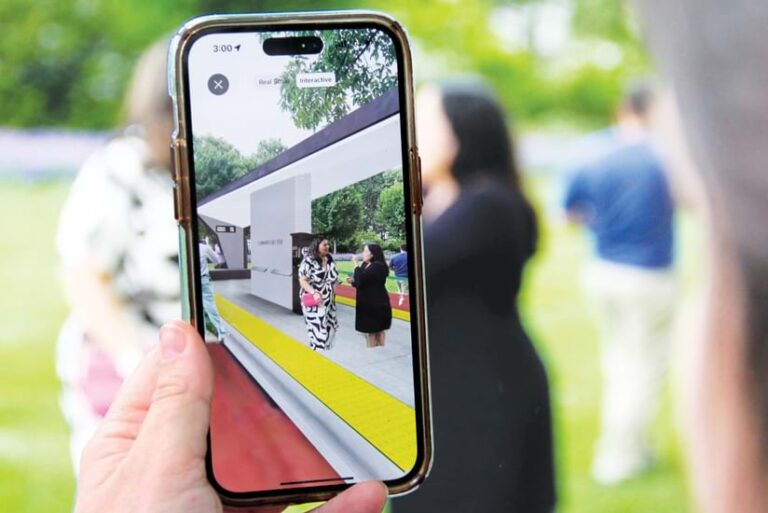 A hand holds a smartphone displaying an augmented reality experience. Two women stand inside a virtual bus station superimposed over a real-world outdoor setting. A hand holds a smartphone displaying an augmented reality experience. Two women stand inside a virtual bus station superimposed over a real-world outdoor setting.