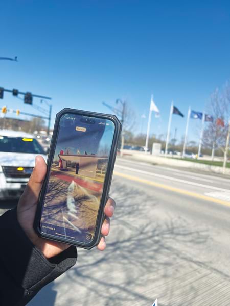 A hand holds up a smartphone displaying an augmented reality-generated transit station in its real-world context. The screen shows a 3D image of a bus station overlaid on the real world as seen through the camera. The background shows a street, trees, and a row of flags. A hand holds up a smartphone displaying an augmented reality-generated transit station in its real-world context. The screen shows a 3D image of a bus station overlaid on the real world as seen through the camera. The background shows a street, trees, and a row of flags.
