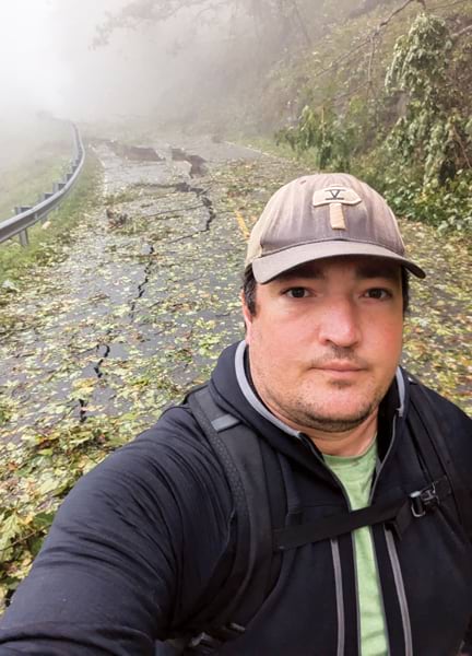 Selfie of a man wearing a baseball cap and backpack on a road through a forest. The road is covered with debris and large cracks from a landslide. The forest is shrouded in fog. Selfie of a man wearing a baseball cap and backpack on a road through a forest. The road is covered with debris and large cracks from a landslide. The forest is shrouded in fog.
