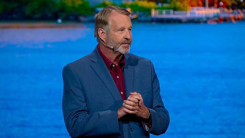 A person wearing a blue suit and clasping their hands in front of a large screen displaying a body of water