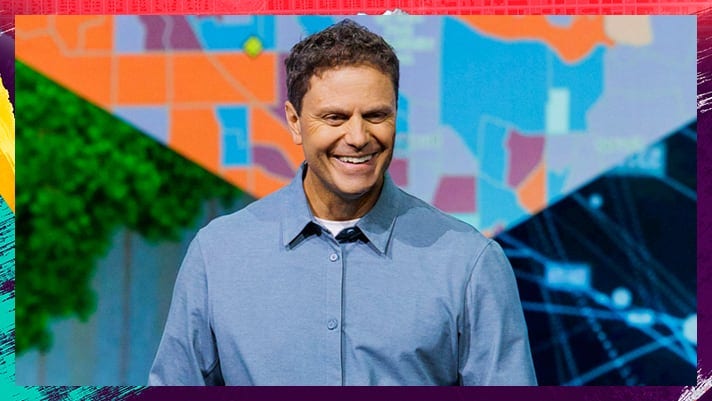 Person standing on stage wearing a blue dress shirt with a large colorful map graphic behind them