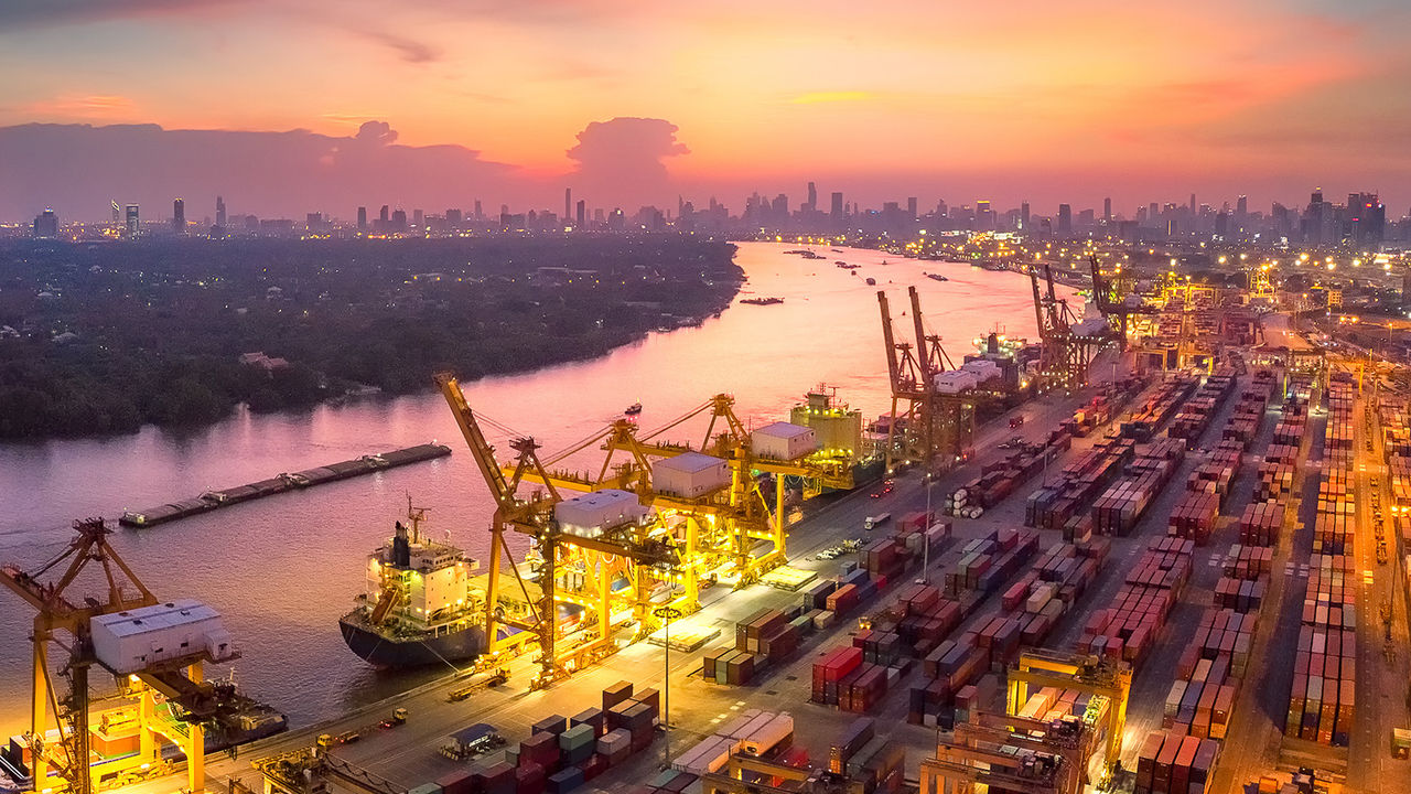 صورة جوية لمنطقة صناعية على رصيف الميناء، ذات إضاءة ساطعة استعدادًا للعمل ليلاً؛ تقع بمحاذاة نهر ساكن، وتظهر أضواء المدينة في المسافة تحت سماء غروب تتدرج ألوانها بين الوردي والبنفسجي