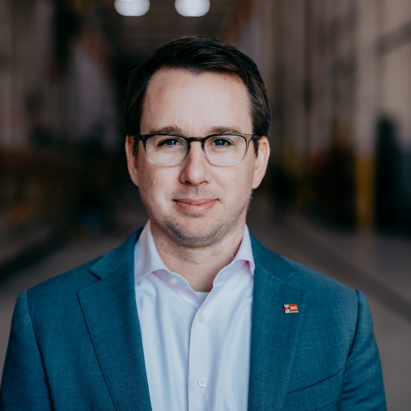 White man with glassess, brown hair, blur blazer light blue collared button down.