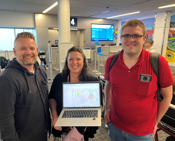 Three people stand at an airport boarding gate. On the left, a man with a beard in a dark gray hoodie smiles toward the camera. In the middle, a smiling woman with long dark hair holds a laptop faced toward the camera; the computer monitor shows a map. On the right, a man with glasses and a red polo shirt smiles towards the camera. He is wearing a backpack and has a cell phone in his front shirt pocket.