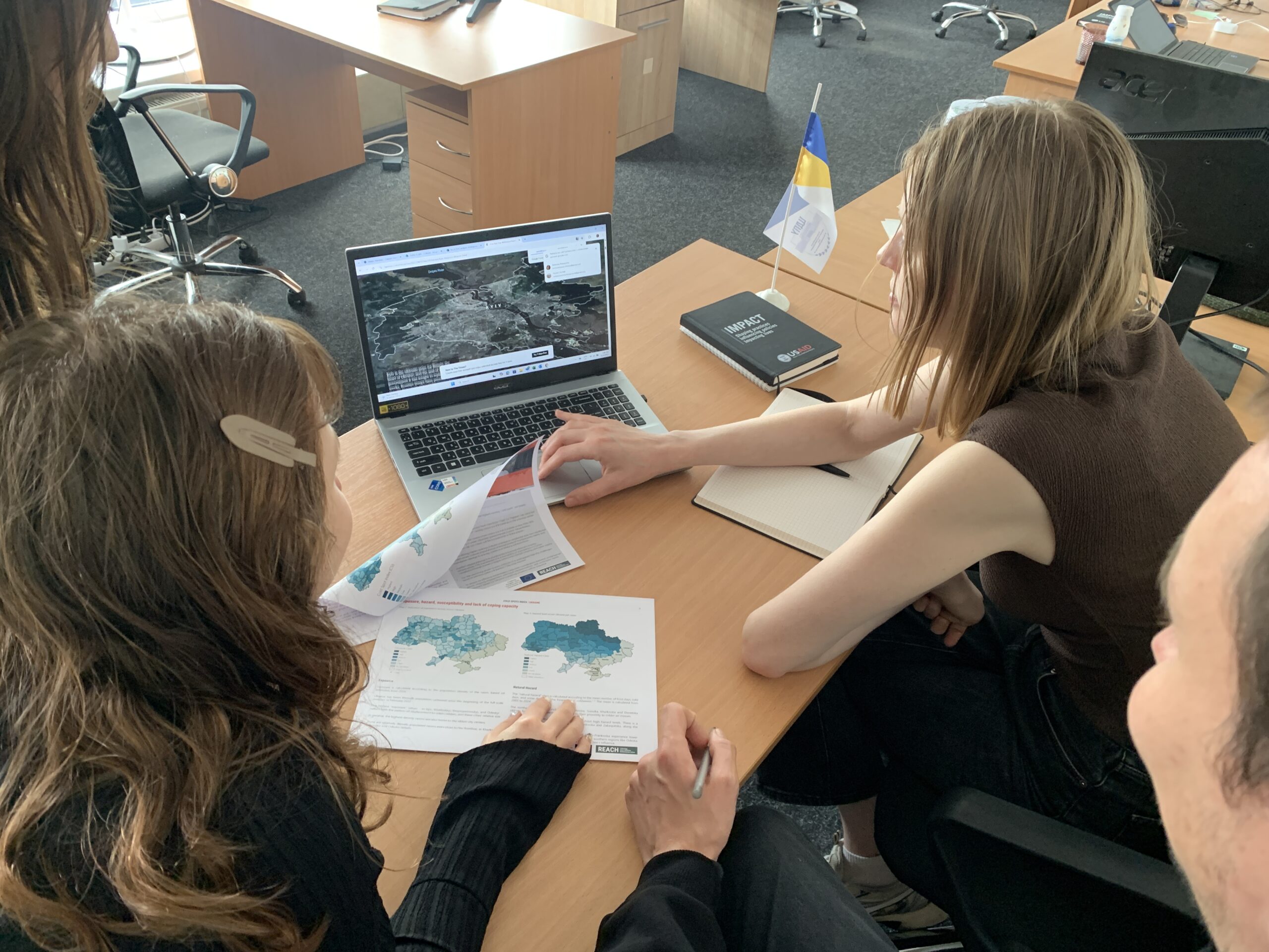 Two women at a desk seen from behind with a laptop open to ArcGIS StoryMaps and a printout of colorful maps they are paging through