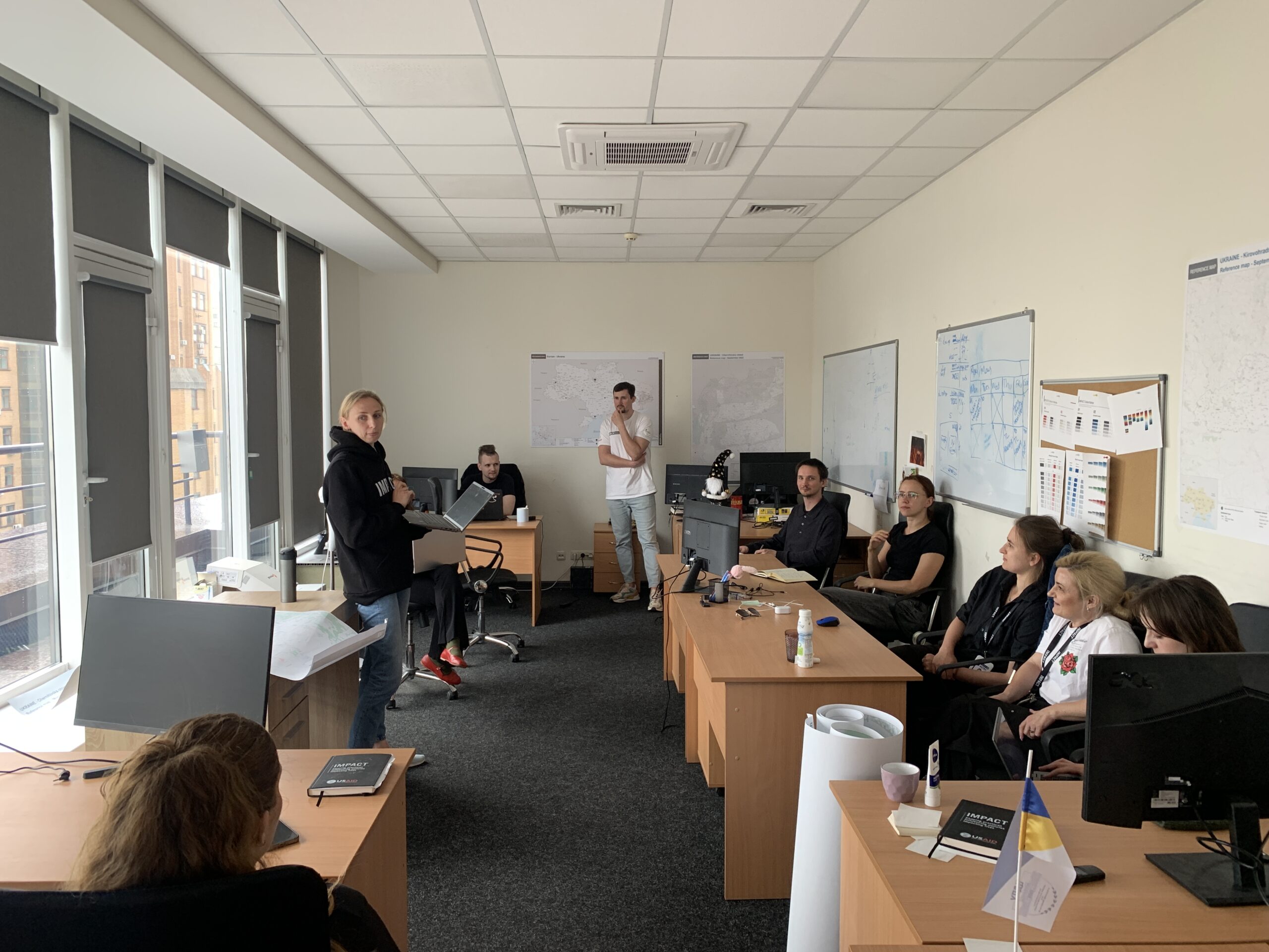 An office with a large group of people casually sitting at desks around the perimeter of the room with the focus on a woman at the front dressed in black casual clothes and a wall of windows behind her