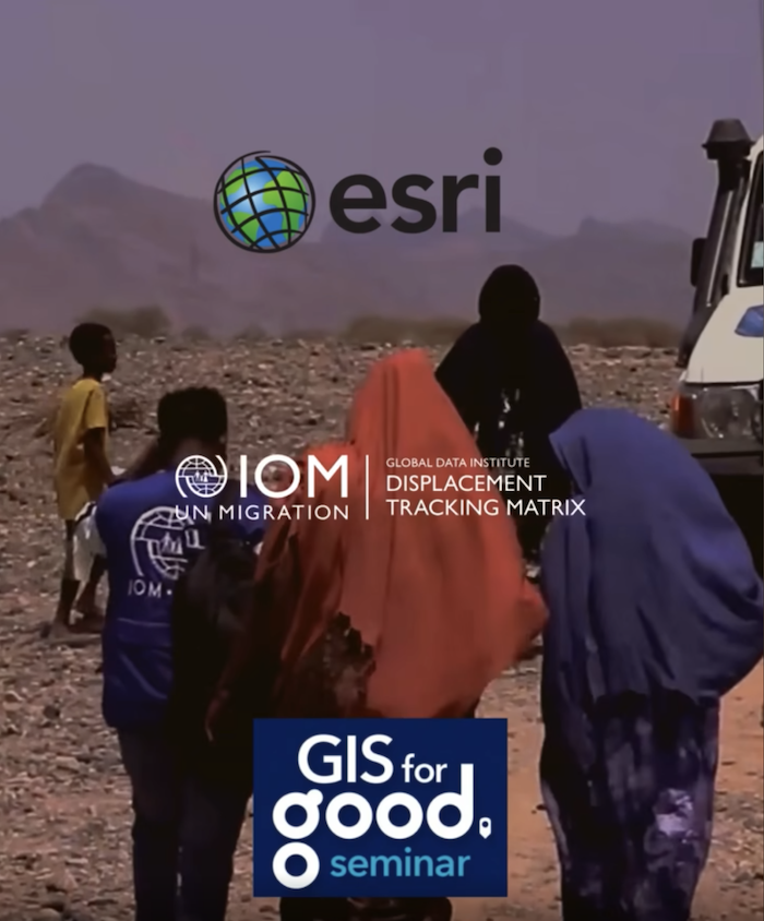 Group of women in traditional clothing walking toward a vehicle parked in a barren, rocky landscape. A man wearing a blue vest with the IOM logo walks with them, all figures facing away from the camera. A child is visible to the left side of the frame also heading towards the vehicle.