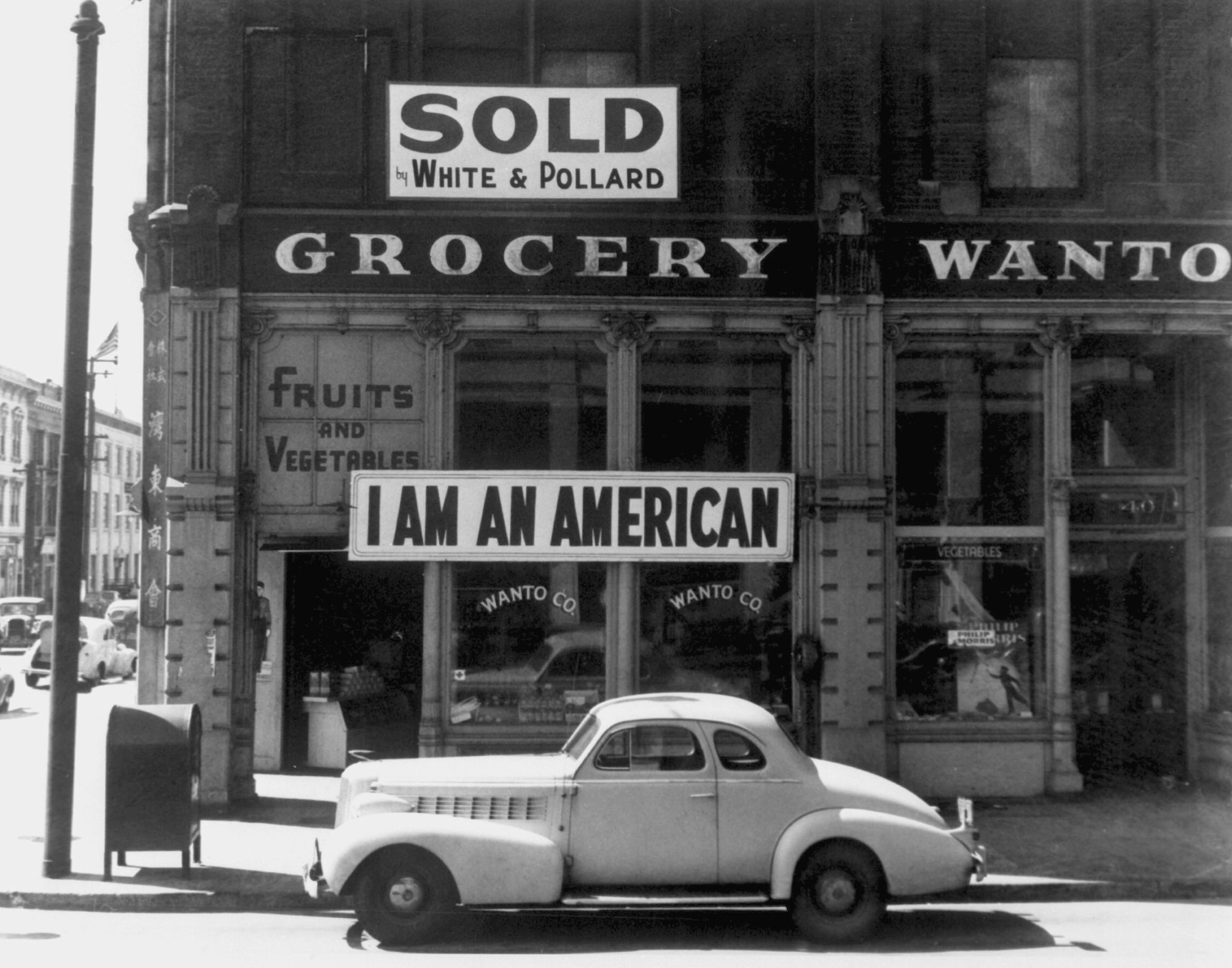 A black and white photograph of a Japanese-owned storefront in California in 1944, with a large banner affixed reading "I AM AN AMERICAN."