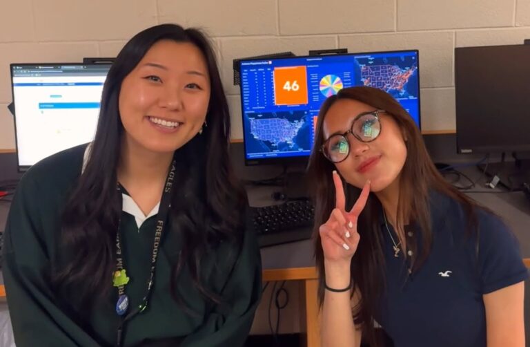 Two women side by side with computer screens displaying a dashboard behind them