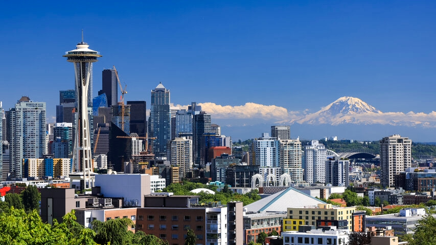 Panoramic view of the Seattle skyline featuring the Space Needle, downtown skyscrapers, and Mount Rainier rising in the distance beneath a clear blue sky.