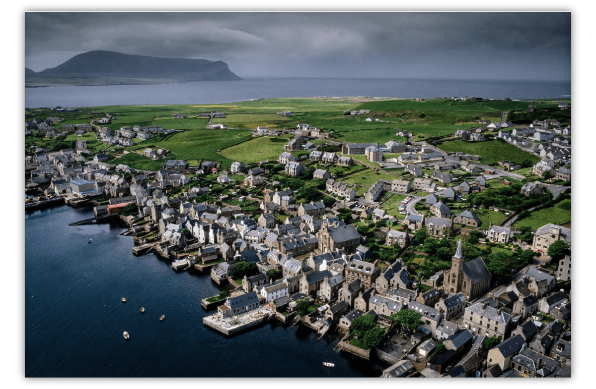 An aerial shot of a historic fishing village showing clustered stone buildings on the waterfront, with expansive green fields behind them An aerial shot of a historic fishing village showing clustered stone buildings on the waterfront, with expansive green fields behind them