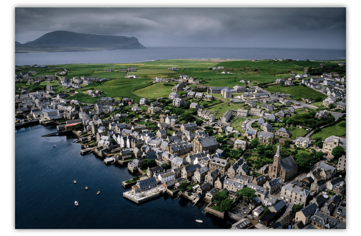 An aerial shot of a historic fishing village showing clustered stone buildings on the waterfront, with expansive green fields behind them