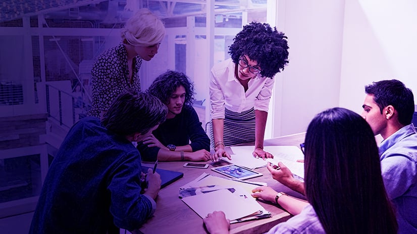 Six people collaborating around a conference room table with papers, folders, tablets, and notepads scattered on the table