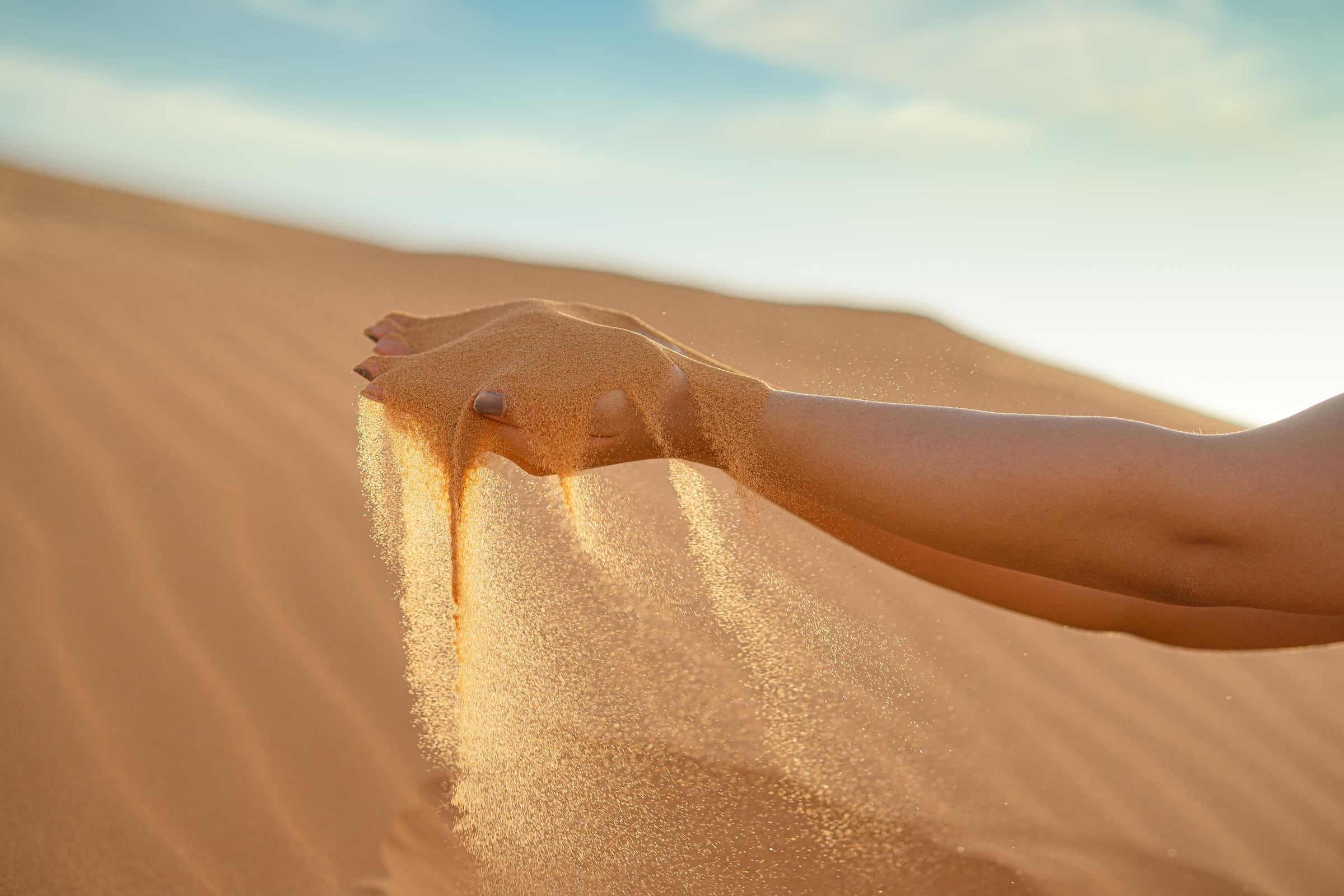 A hand at the end of an extended arm holds a pile of fine sand which is falling from the hand. A sand dune is in the background