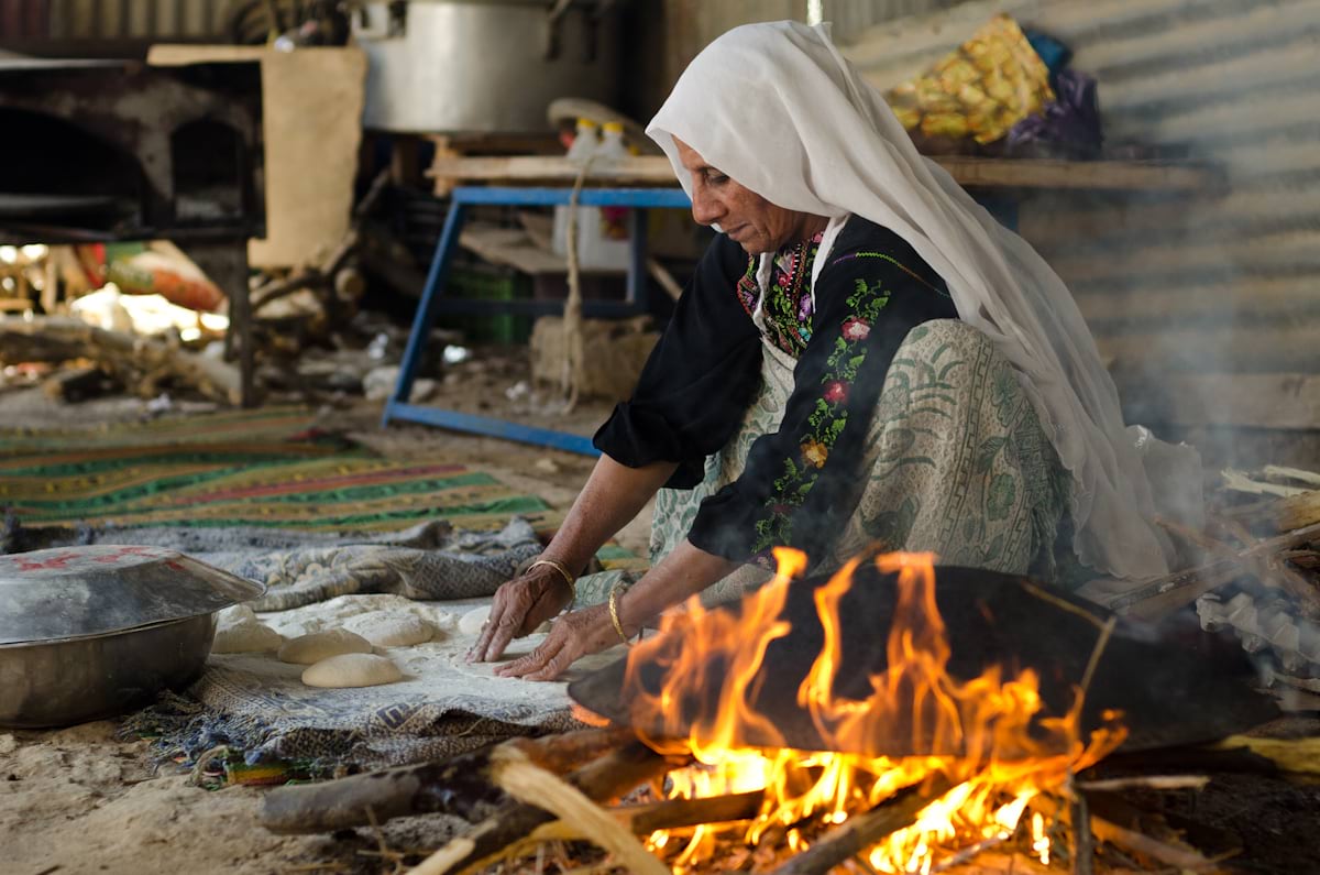 A person rolls bread dough on the ground by an open fire